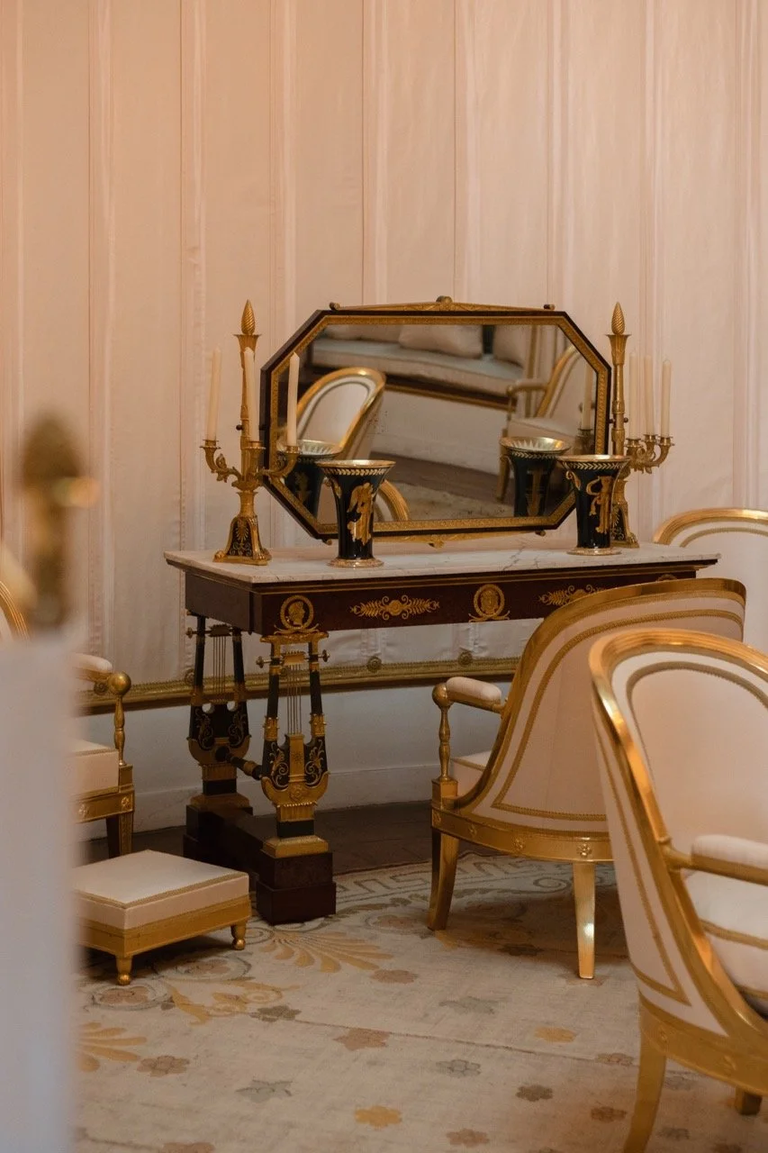 An elegant dressing table with a large mirror, decorated in black and gold with candelabras and small cups, surrounded by ornate chairs in a luxurious room with light-colored wood-paneled walls and a patterned rug.