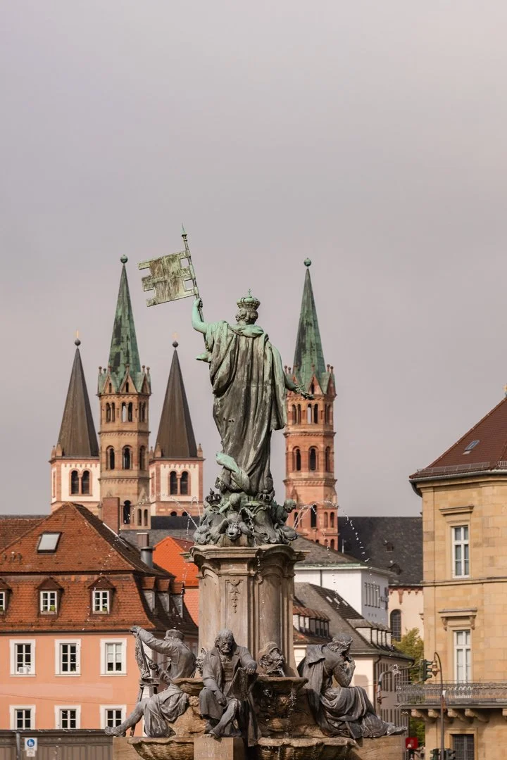 A historic fountain featuring a statue of a crowned woman holding a flag, with water spouting from the base, surrounded by a cityscape with church steeples and historic buildings in the background.