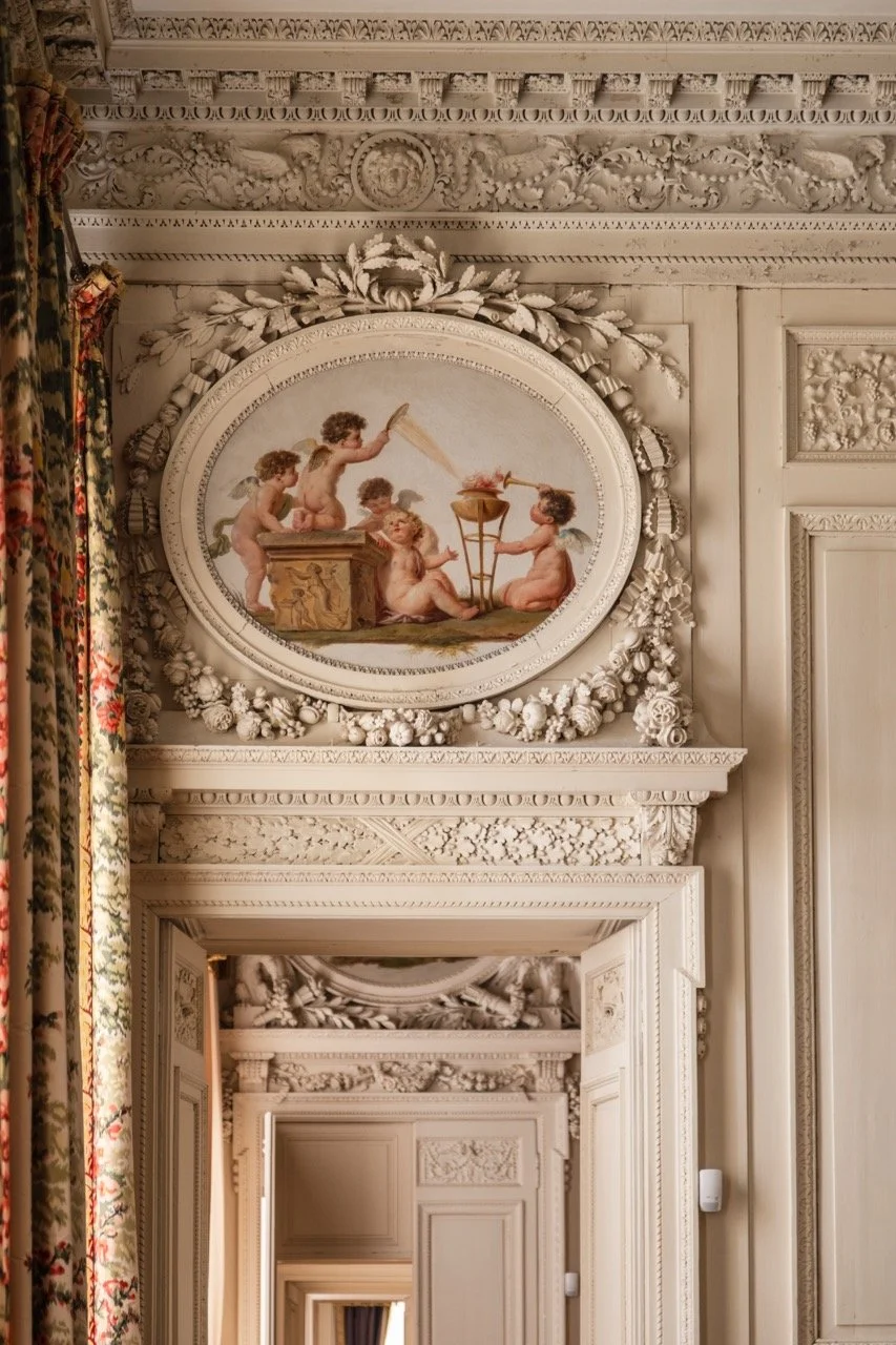 Ornate interior with elaborate white molding and a large oval painting of cherubs, some with wings, surrounding a small fire, surrounded by a decorative floral and scroll border.