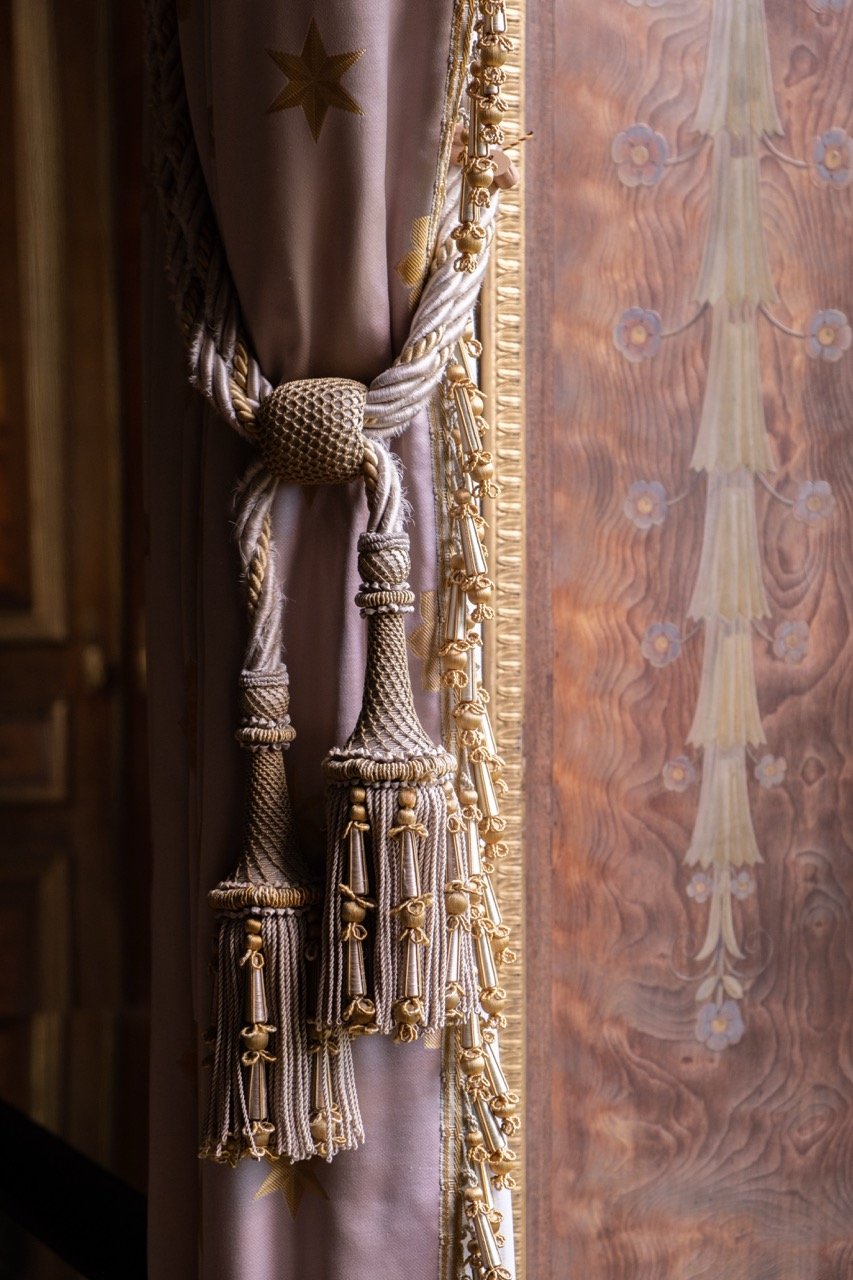 Close-up of ornate curtain tieback with gold accents, draped over a curtain with gold star embroidery, next to a patterned wooden wall with floral designs.
