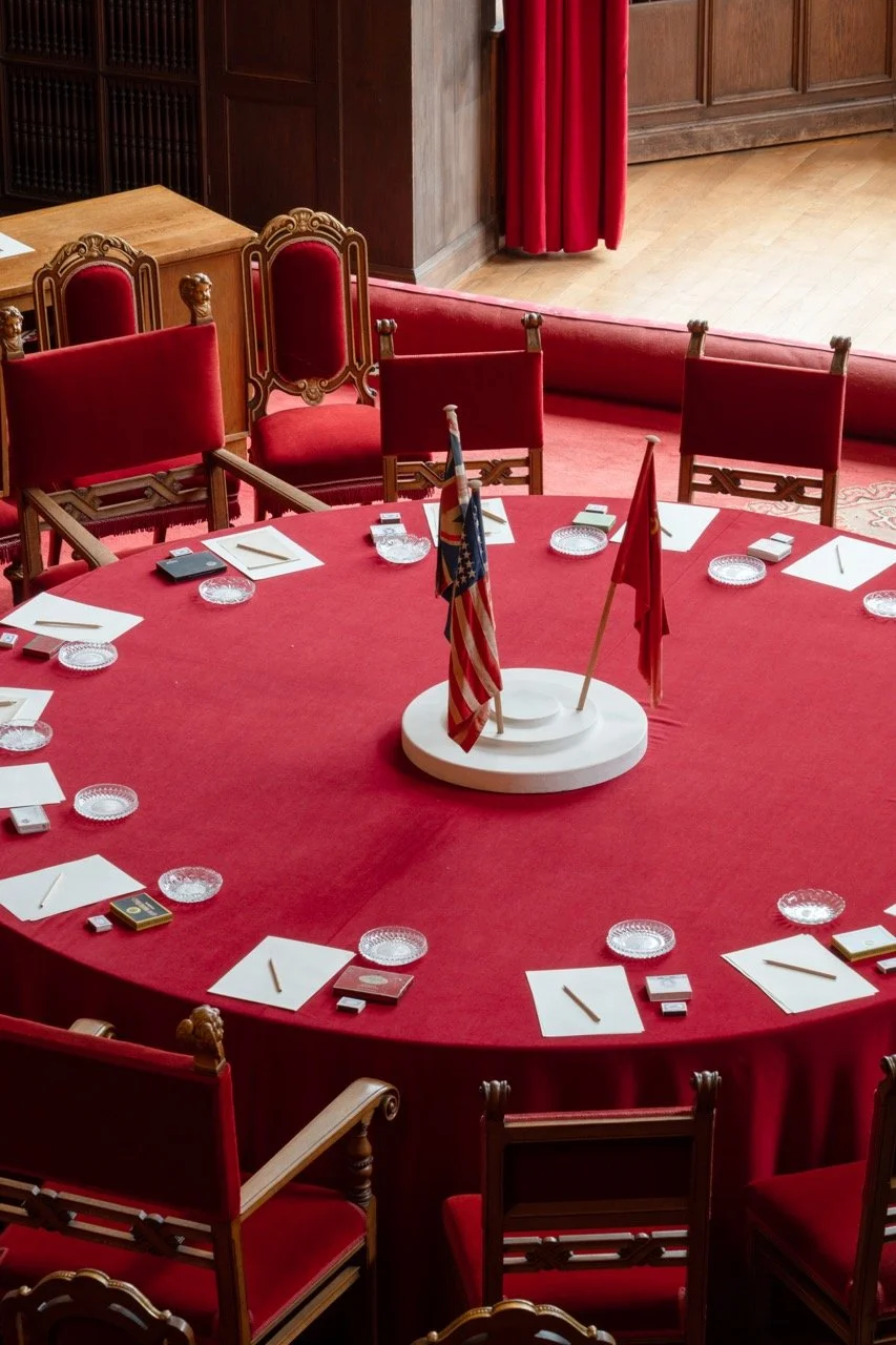 Round table with red tablecloth, eight chairs, and flags in the center, set for a formal meeting or event, with white paper and pens for each seat.