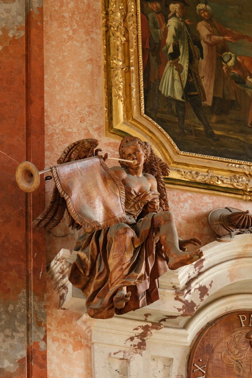 Baroque wooden sculpture of a seated woman playing a pipe, located inside an ornate room with a large framed painting on the wall above.
