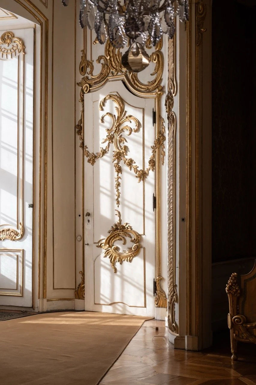 An ornate white door with gold embellishments and intricate carvings, casting shadows on a beige floor in a luxurious room with a chandelier overhead.