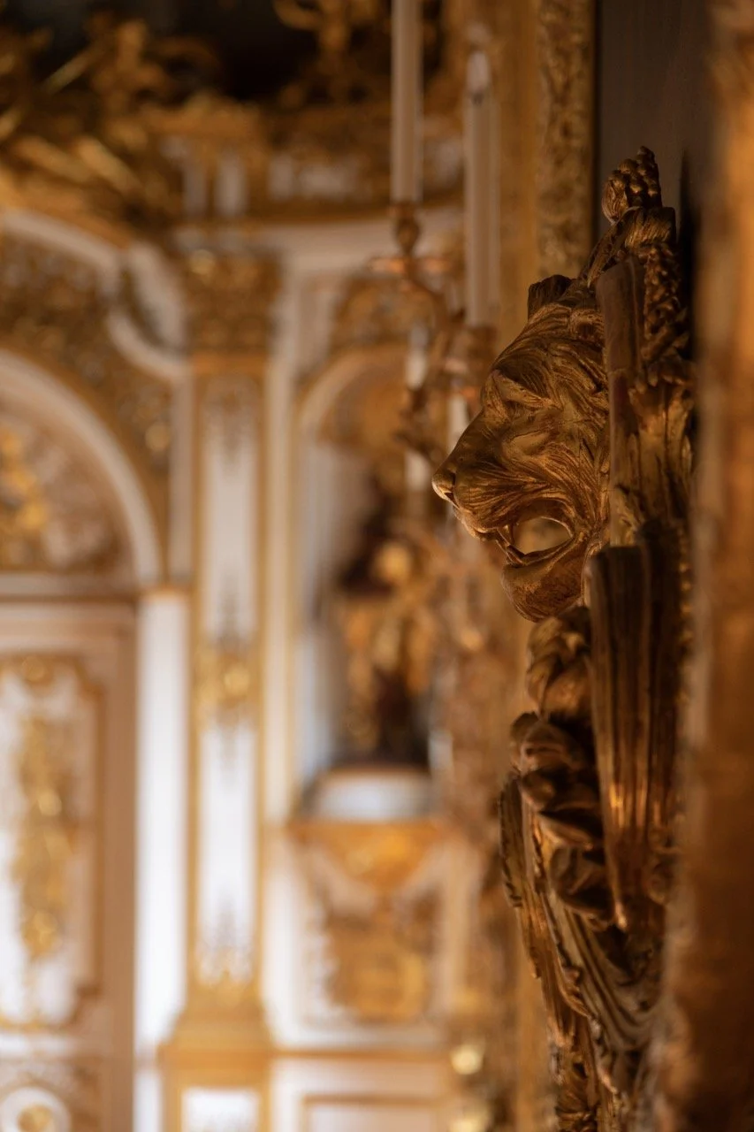 Close-up of a gilded gold carved sculpture of a lion's head on a decorative wall in a grand, ornate room with gilded details and classical architecture.