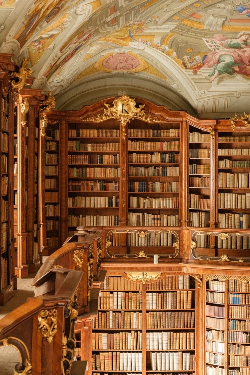 An ornate, historic library with tall wooden bookshelves filled with old books, detailed gold embellishments, and a painted ceiling featuring classical artwork.