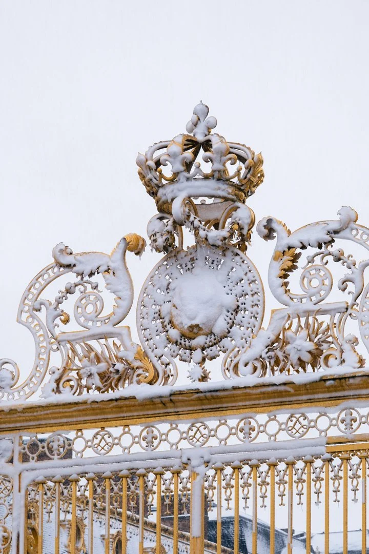 Ornate golden and white wrought iron gate with clock and crown, covered in snow