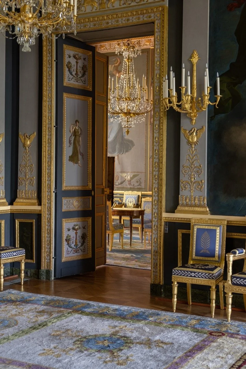 An ornately decorated room with gold and blue accents, featuring chandeliers, gilded wall sconces, classical artwork, and elegant furniture.