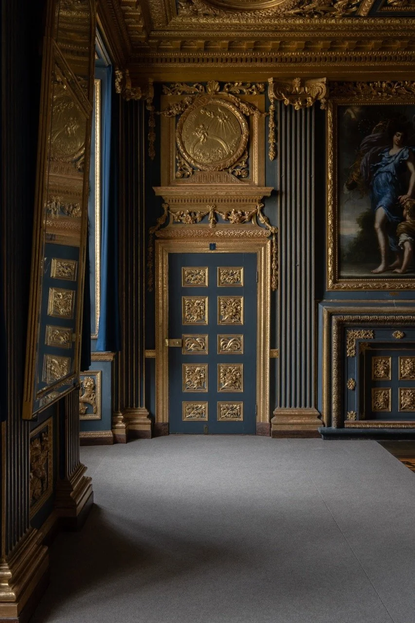 Interior of a room with ornate blue and gold decor, a decorated door, a large framed painting of a woman, and gold embellishments on walls and ceiling.
