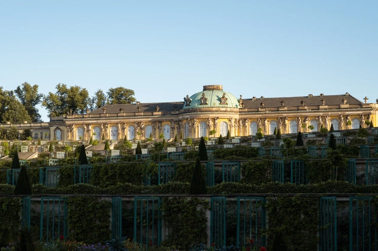 A grand historic building with a domed roof, classical architecture, and garden terraces with green shrubbery and trees.