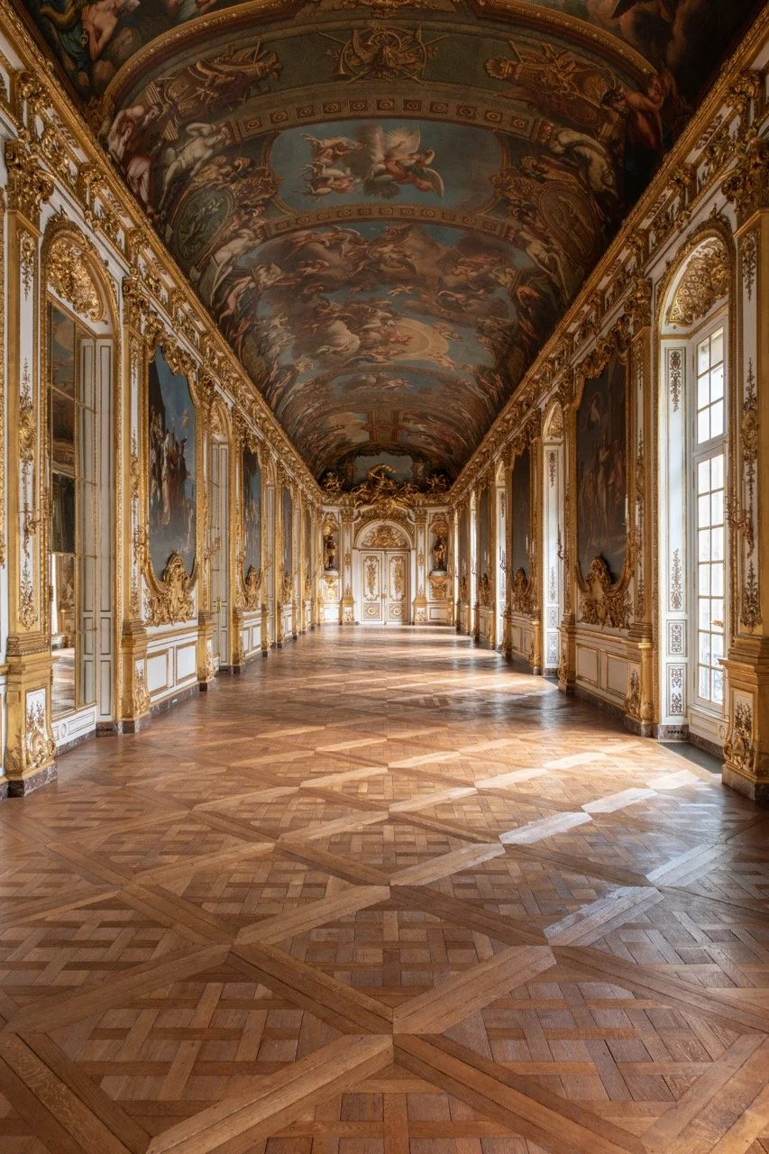 An ornate, gilded hall with intricate gold framing, large windows, and a painted ceiling with classical artwork, featuring a polished wooden parquet floor.