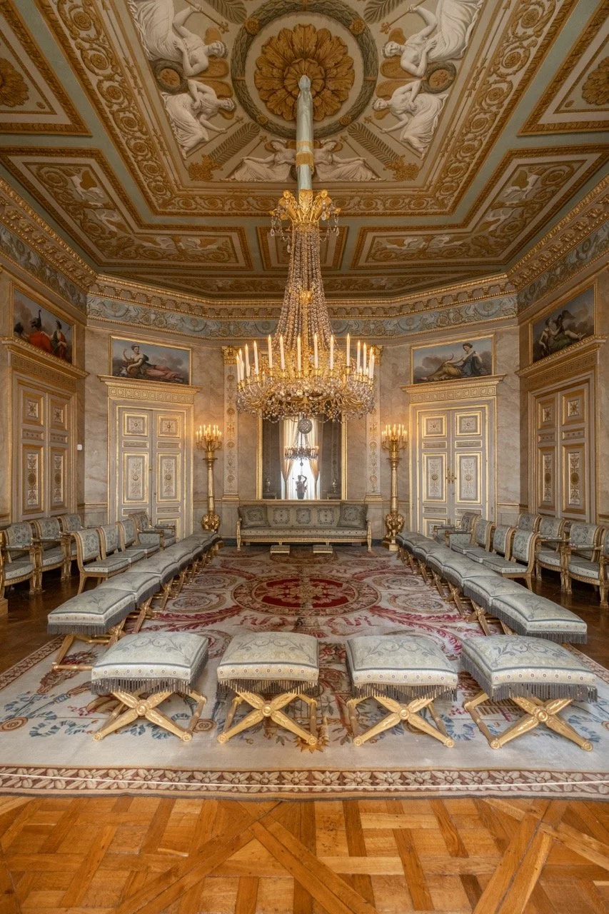 A grand, opulent living room with gold-accented ornate furniture arranged in a U-shape, a large chandelier hanging from a decorated ceiling, and framed classical paintings on the walls.