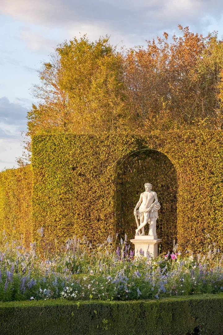 A marble sculpture of a man stands on a pedestal in a garden with flowering plants, framed by an arched bush, with tall trees and a cloudy sky in the background.