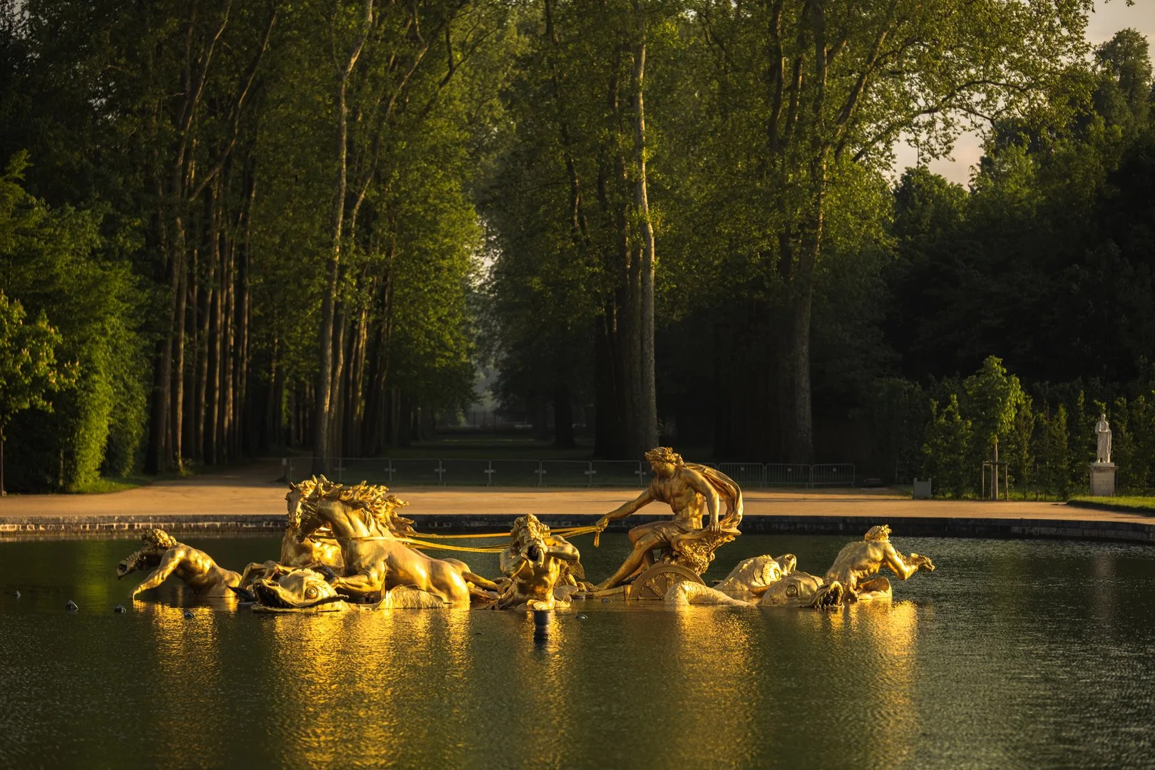 Golden fountain sculpture depicting a chariot pulled by horses and a figure holding reins, in a pond with trees in the background.