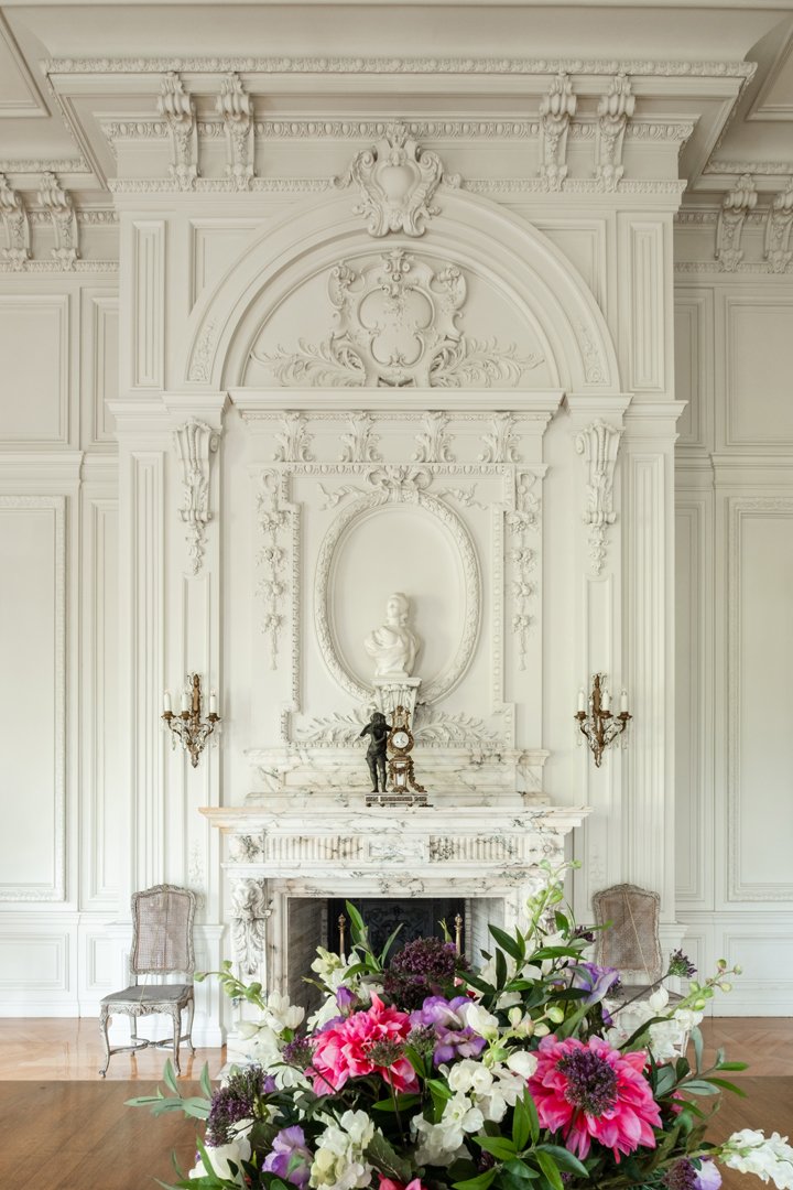 Elegant white room with ornate detailing, a marble fireplace, two antique chairs, a classical bust, a clock, and a flower arrangement in the foreground.