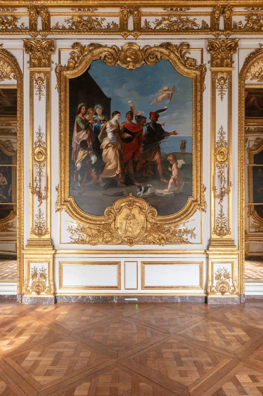 A large classical painting depicting a group of mythological or historical figures, set in ornate gold and white frame in a grand room with parquet wood floors.