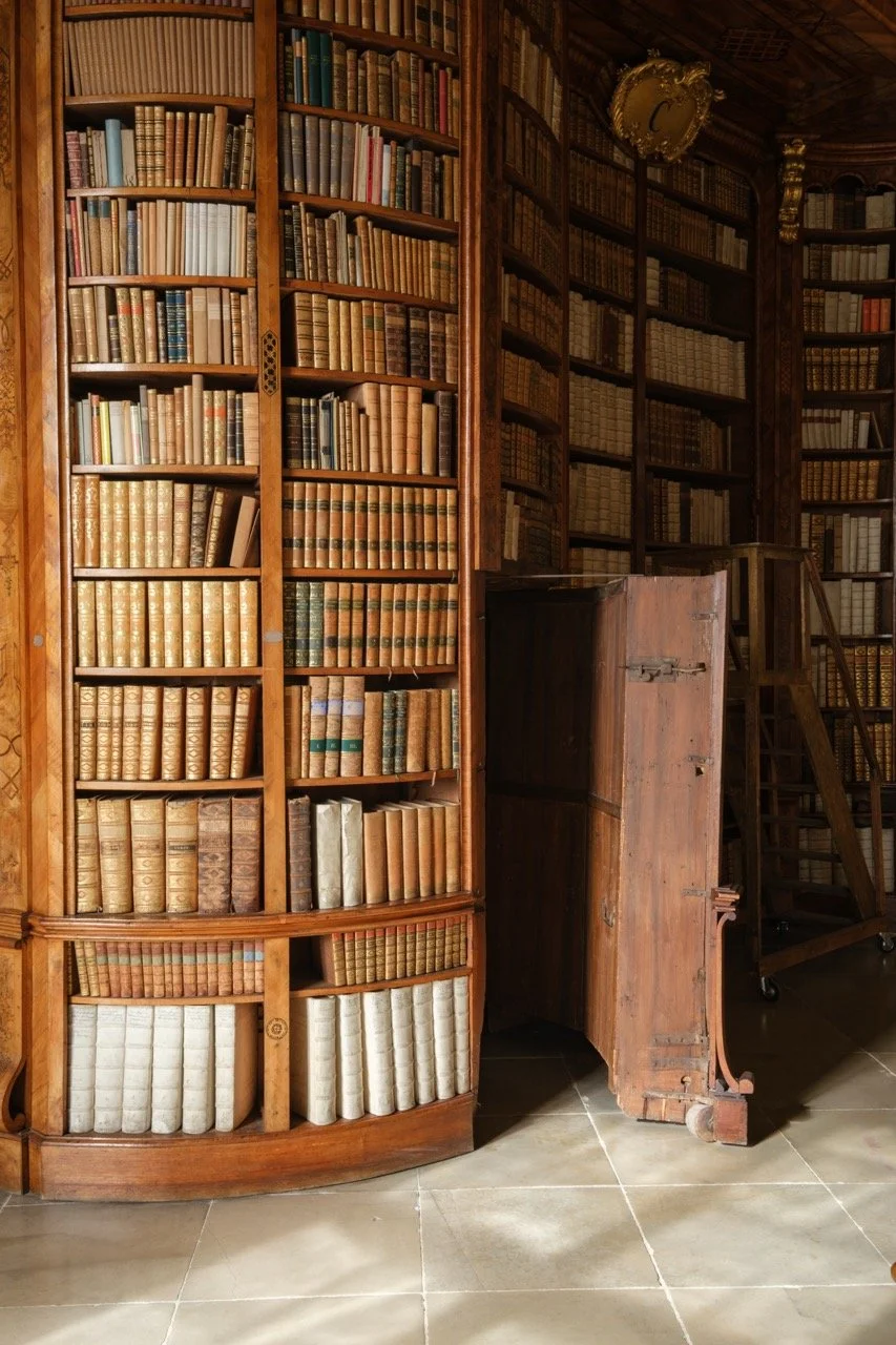 A large wooden bookcase filled with old books in a library or study, with some books open and a small wooden cabinet on a tiled floor.