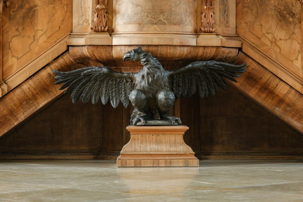 Bronze sculpture of a mythical griffin with outstretched wings on a wooden pedestal, situated in a grand indoor space with wood-paneled walls.