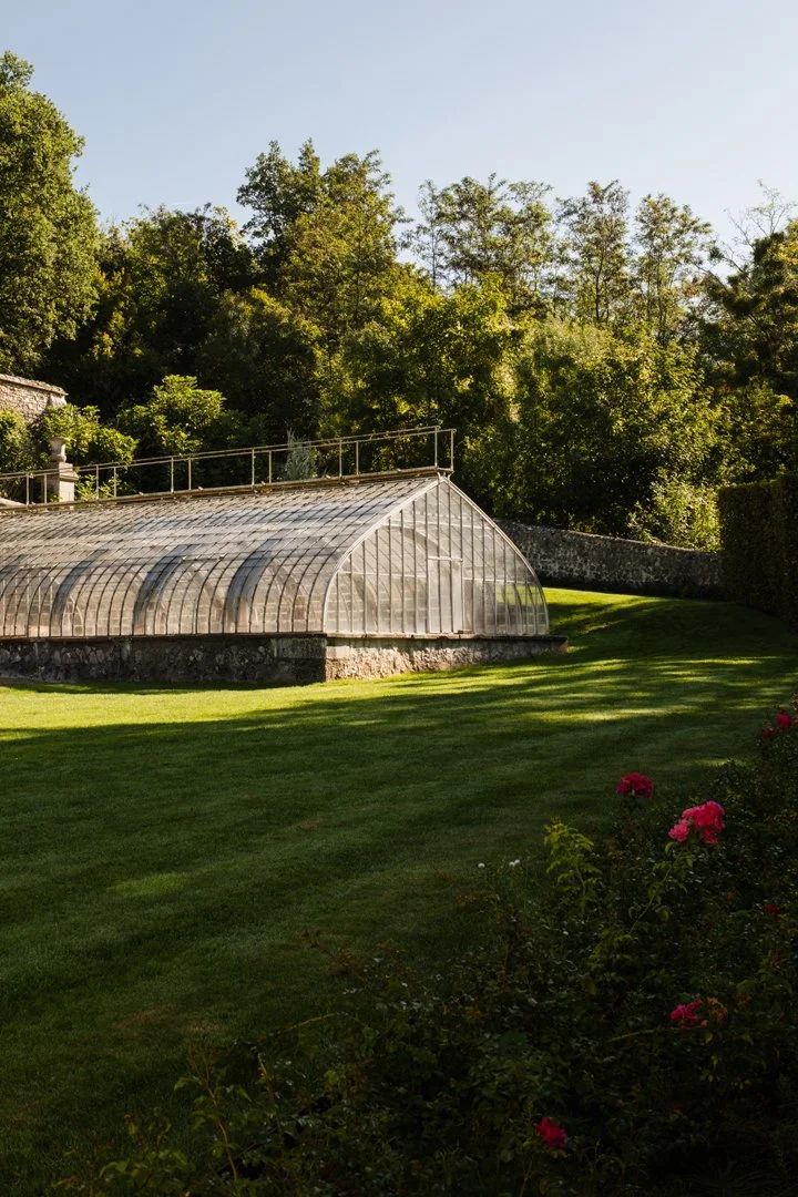 A glass greenhouse with a rounded roof in a lush garden, surrounded by green grass and trees.