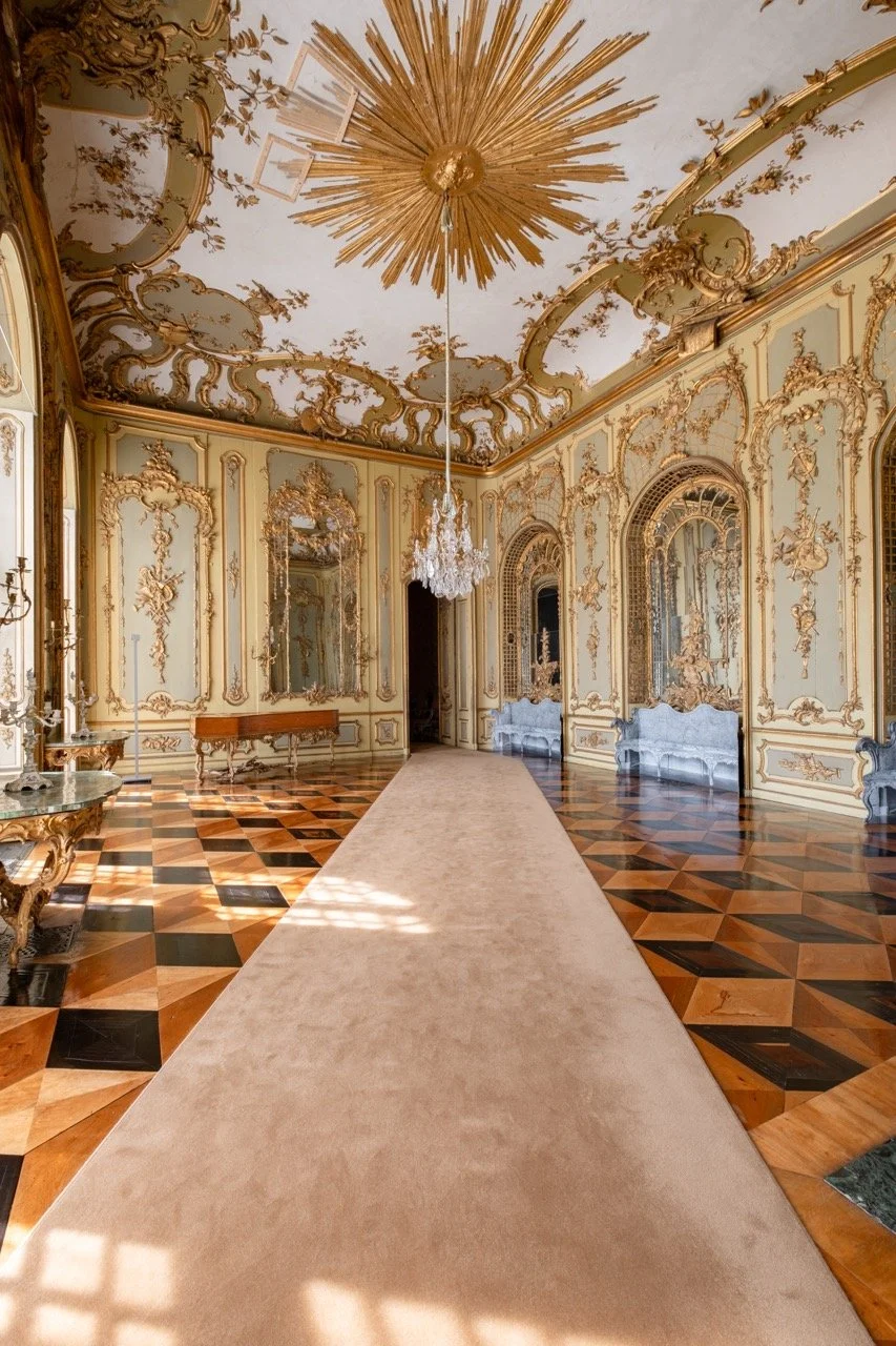 Luxurious palace interior with ornate gold detailing, elaborate moldings, and large mirrors, featuring a high ceiling with gold sunburst design, crystal chandelier, polished wooden parquet flooring, and a beige runner carpet.