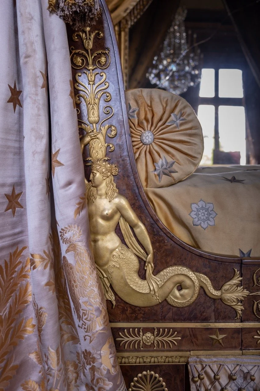 Close-up of an ornate antique bed with gilded lion and fish figurehead, intricate wood carvings, gold-embroidered bedding, and luxurious curtains, with a window and chandelier in the background.