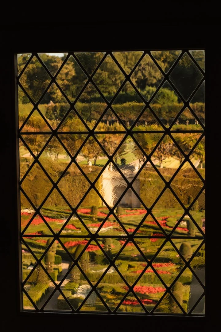 View through a decorative window with diamond-shaped patterns showing a garden with a water fountain, green grass, and pink flowers.