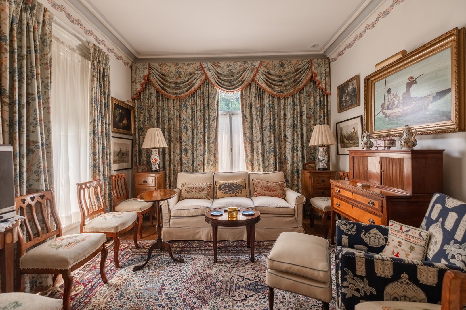 Living room with floral curtains, wooden furniture, a white sofa with cushions, a blue armchair with cushions, and a patterned rug.