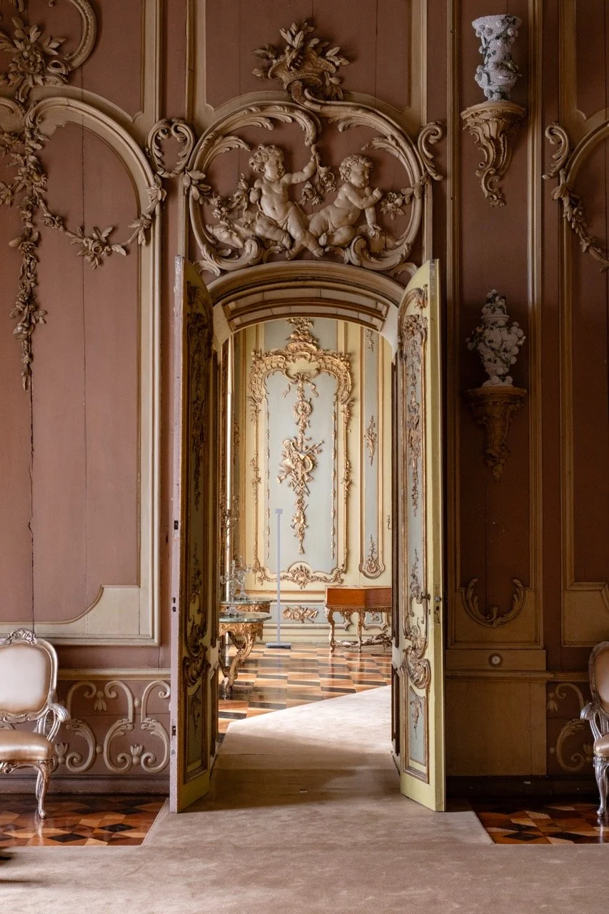 Opulent room with ornate gold and pastel decor, intricate wall carvings, and a pair of matching chairs along the walls, viewed through an open decorated door.