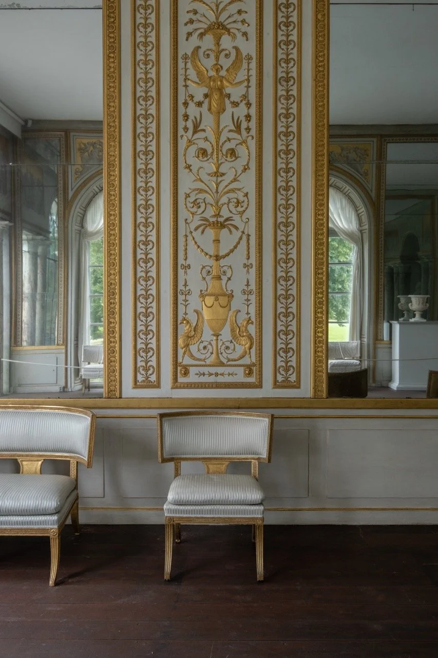A room with ornate gold and white wall decorations, including a large panel with intricate gilded designs and two chairs with white upholstery and gold trim in front of the wall.