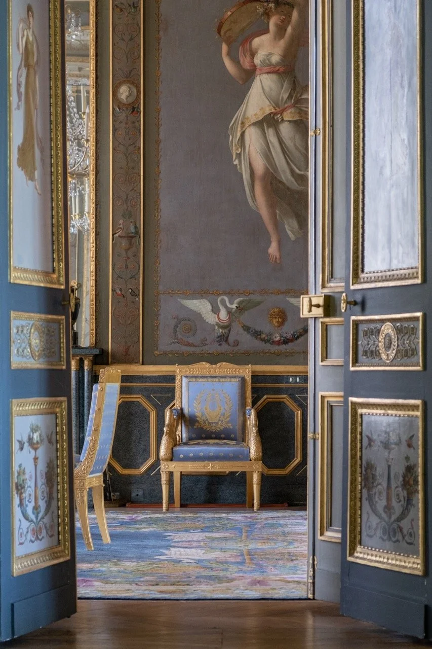 View of an elegant, ornate room decorated with gold accents, featuring a large classical painting of a woman holding a shield, with detailed floral and mythological motifs, seen through open painted doors.