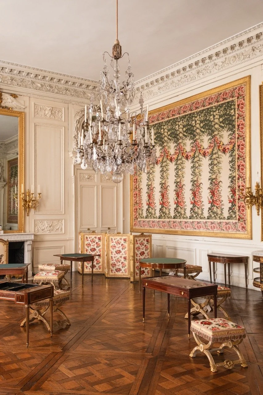 An elegant room with ornate white walls, a large crystal chandelier hanging from the ceiling, a floral tapestry on the wall, and antique furniture including tables and chairs with floral upholstery, all on a polished wooden floor.