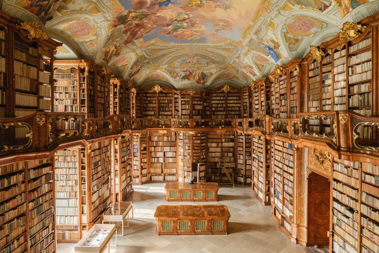 A grand, historic library with tall, wooden bookshelves lining the walls and filling the space with books. The ceiling is decorated with elaborate fresco art featuring angels and clouds. A large wooden table is positioned in the center of the room, i