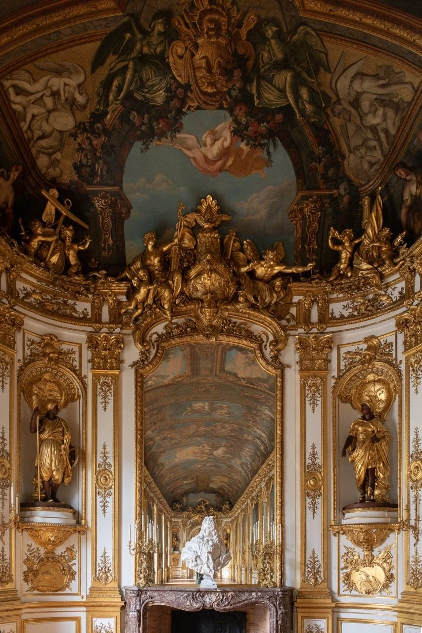 Baroque interior with gold ornate decorations, statues, and a large mirror reflecting a painted ceiling, with a white marble sculpture at the center.