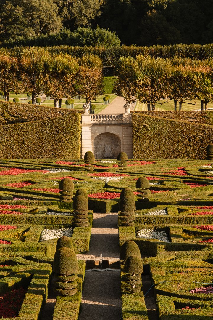 A beautifully landscaped formal garden with trimmed hedges, topiary, and flower beds, featuring a pathway and a small fountain, with trees and shrubs in the background.