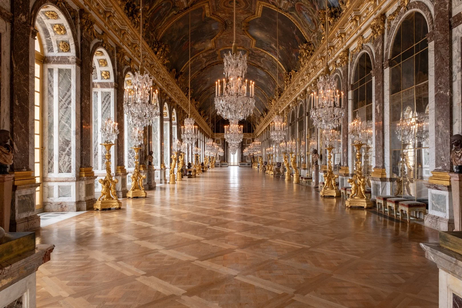 A grand, ornate hall with a high, arched ceiling decorated with gold accents and chandeliers. The hall features tall windows along the sides, and is lined with marble columns and gold sculptures. The floor is made of polished wood arranged in a herri
