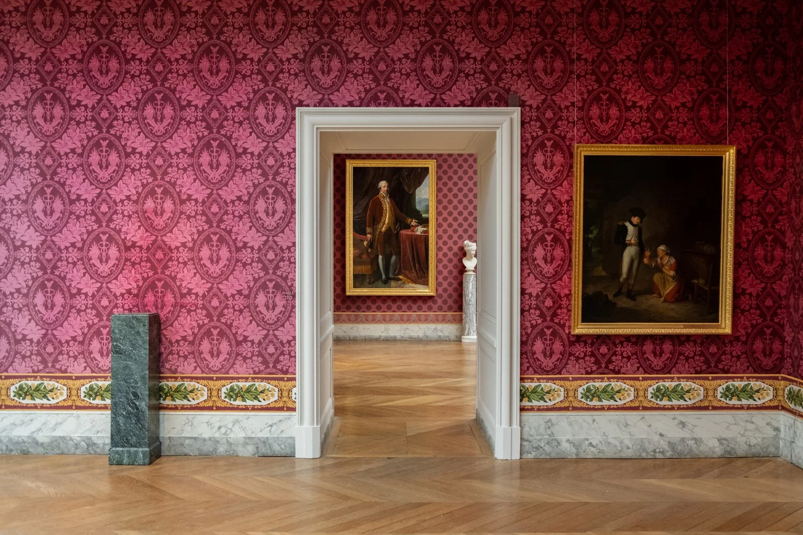 Interior of an art gallery with pink patterned wallpaper, two framed oil paintings, and a marble floor. The first painting is a portrait of a man in historical clothing, and the second painting depicts two figures, one standing and one kneeling, in a
