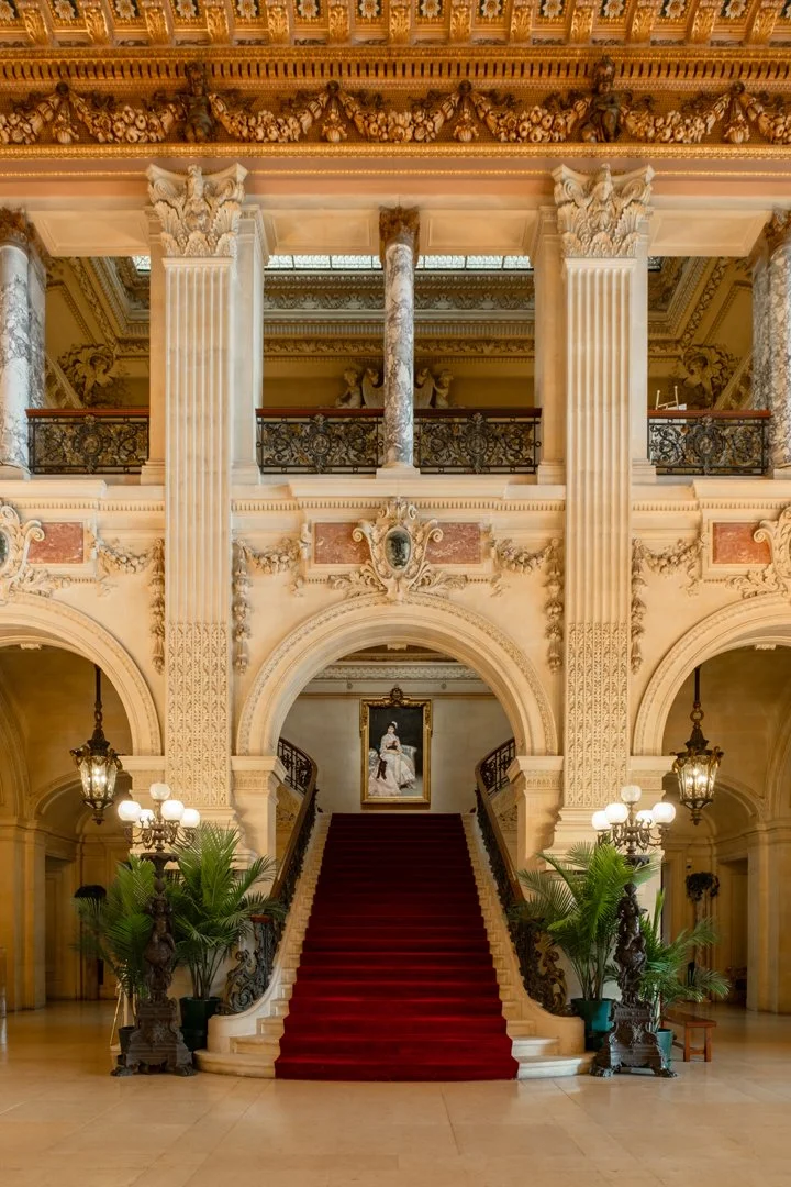 Grand staircase with a red carpet in a luxurious, ornate hall featuring marble columns, decorative moldings, chandeliers, and classical paintings.
