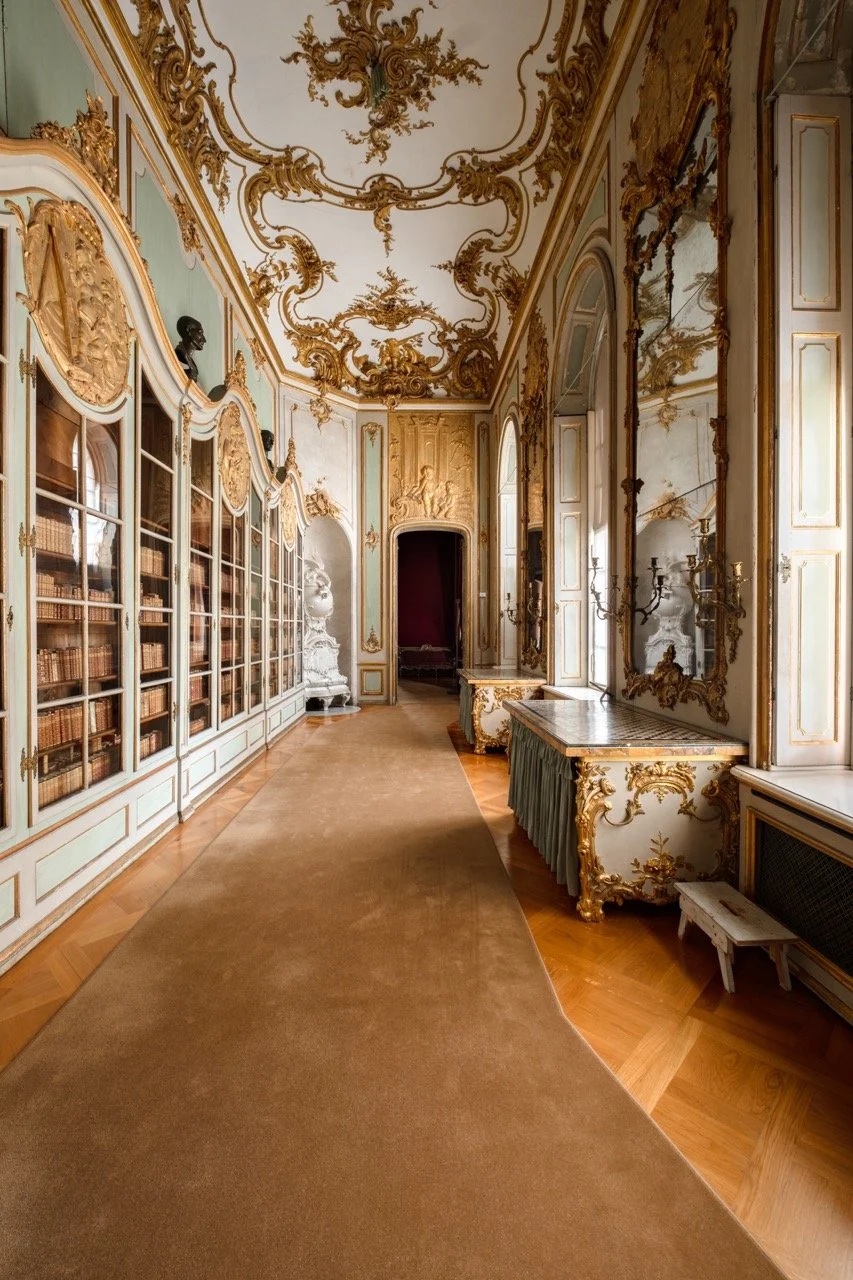 An ornate, classical library room with gold gilded decorations, large mirrors, and a row of bookshelves filled with books.