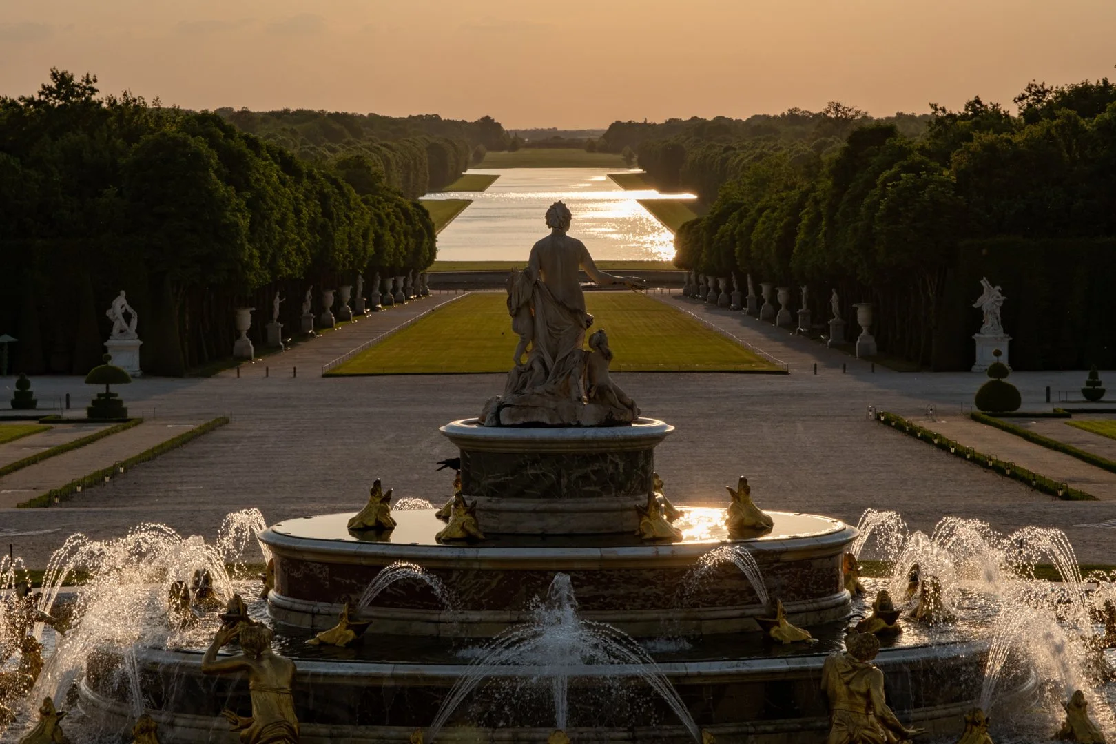 A grand classical-style fountain with statues, in a garden with symmetrical trees and pathways, overlooking a canal at sunset.