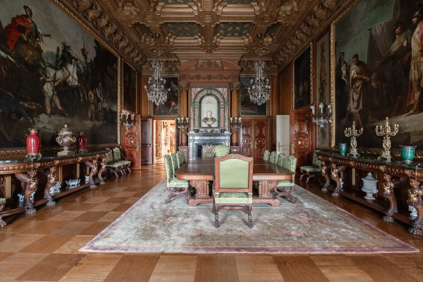 A luxurious, ornate dining room with a large wooden table and green upholstered chairs. The room features elaborate wooden paneling, chandeliers, and large classical paintings on the walls. A fireplace with decorative busts and statues is at the back