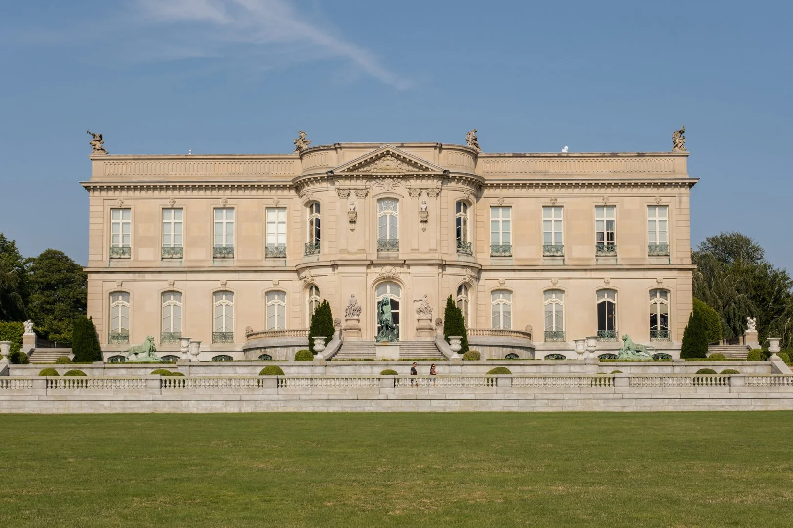 A grand, historic palace with beige stone walls, multiple windows, and ornate sculptures on the roof and facade, surrounded by a manicured lawn and trees.
