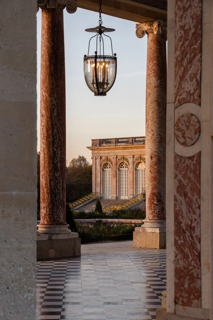 View through an archway with red marble columns showing a historic building with large windows and an ornate staircase in the background. A hanging lantern is visible in the foreground.
