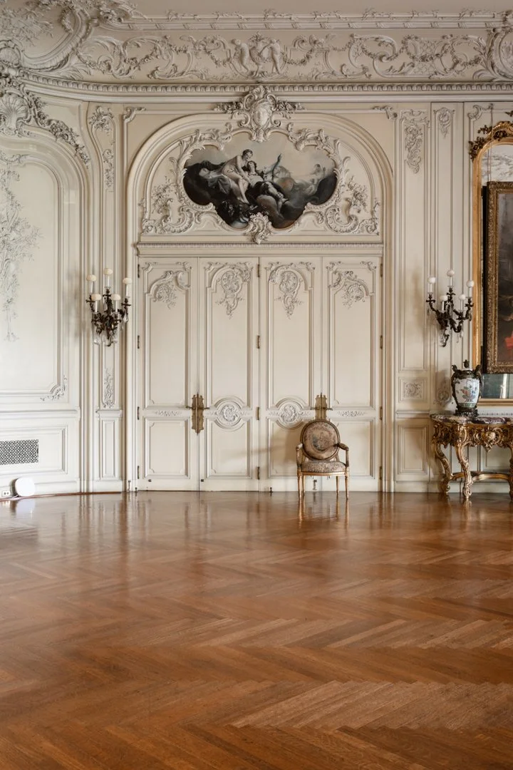 An elegant, ornate room with white decorative molding, gold accents, and a painting above double doors. There are wall sconces with candles, a small upholstered chair, and a wooden table with a decorative vase. The wooden floor has a herringbone patt