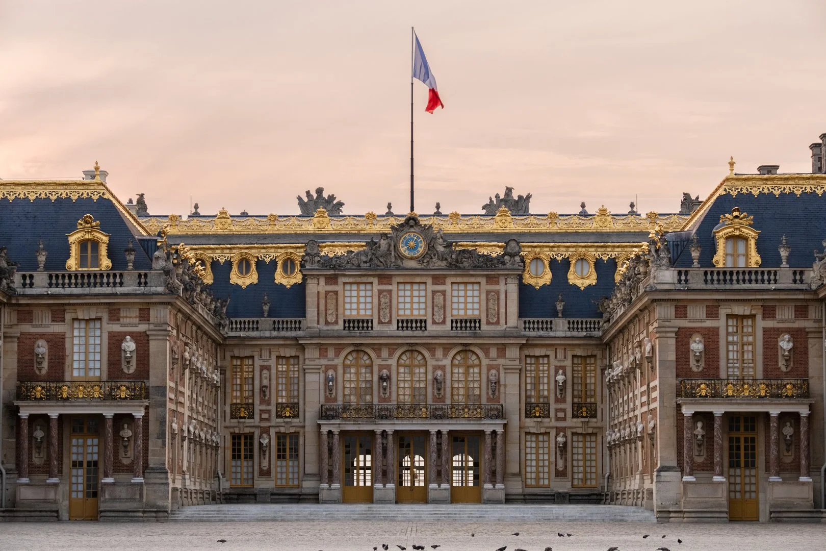 The Palace of Versailles in France showing an elegant historic building with gold accents, sculptures, large windows, and a French flag flying on top in the evening sky.