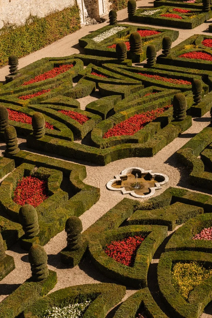 A meticulously maintained formal garden with trimmed hedges, colorful flower beds, topiary trees, and a central fountain.