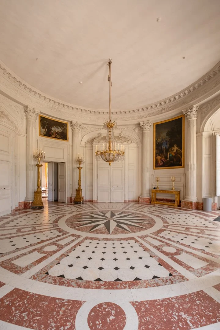 Elegant, ornate room with a circular, patterned marble floor, a central chandelier, white paneled walls with gold accents, tall Corinthian columns, and large framed paintings.