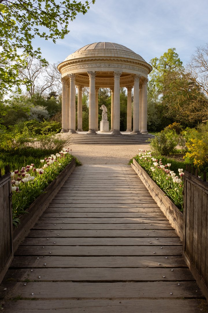 A wooden pathway leading to a neoclassical pavilion with columns and a domed roof, surrounded by lush greenery and blooming flowers.