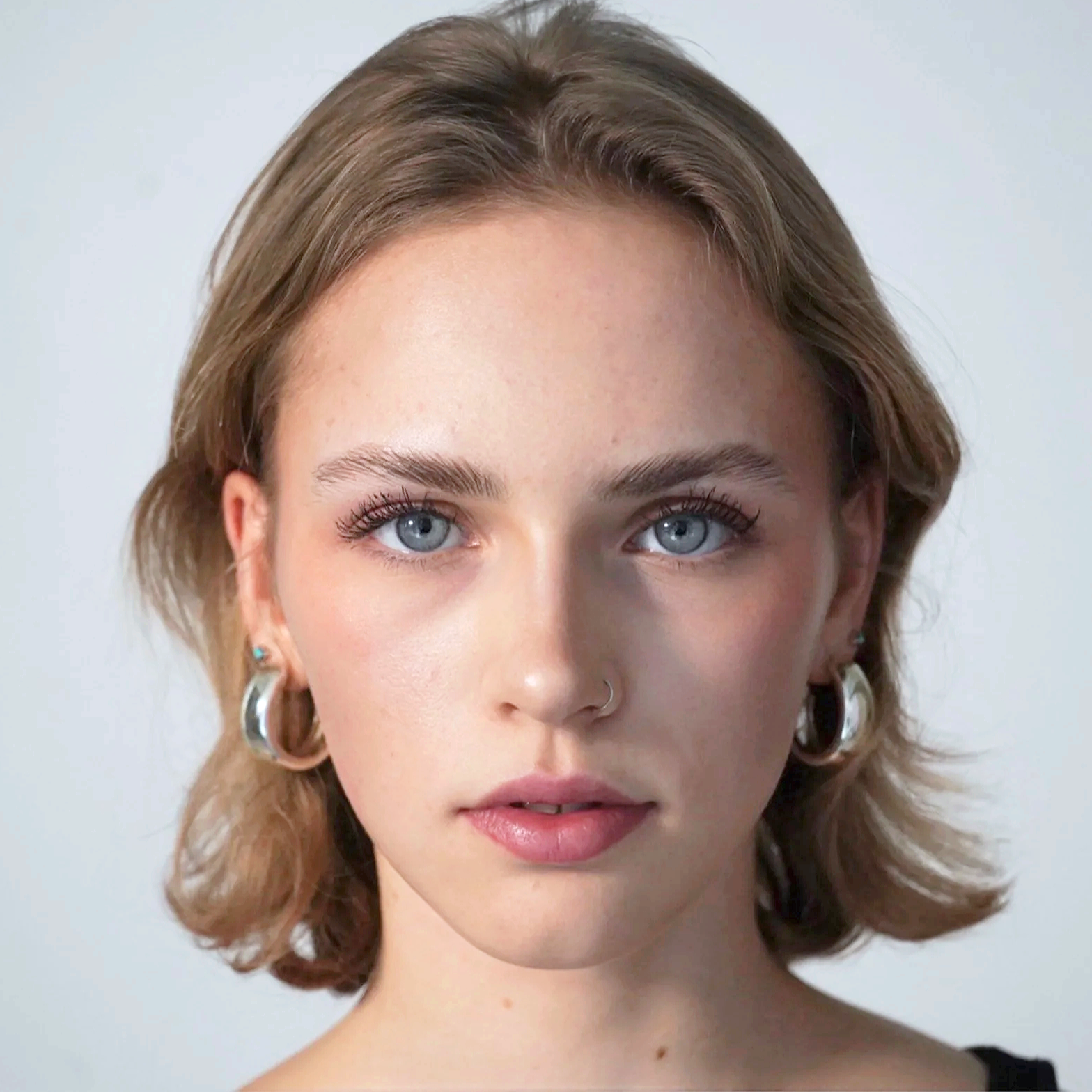 Nahaufnahme eines jungen Mädchens mit blauen Augen, kurzen braunen Haaren, Nasenpiercing und Ohrringen, vor einem neutralen Hintergrund.