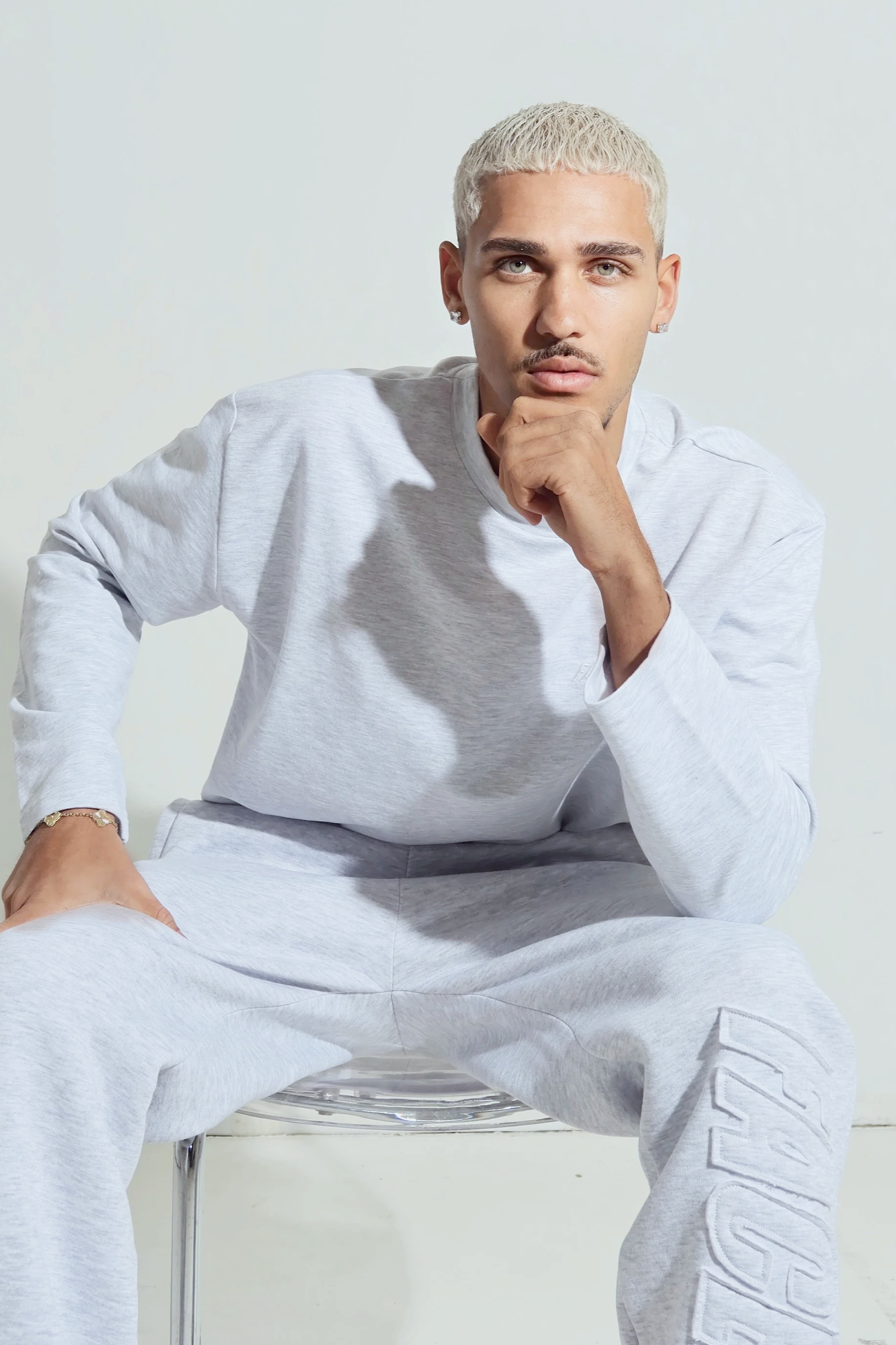 Junge Mann mit blonder Kurzhaarfrisur, grauem Jogginganzug, sitzend auf einem Stuhl, blickt ernst in die Kamera, Hand am Kinn, helles Hintergrund.