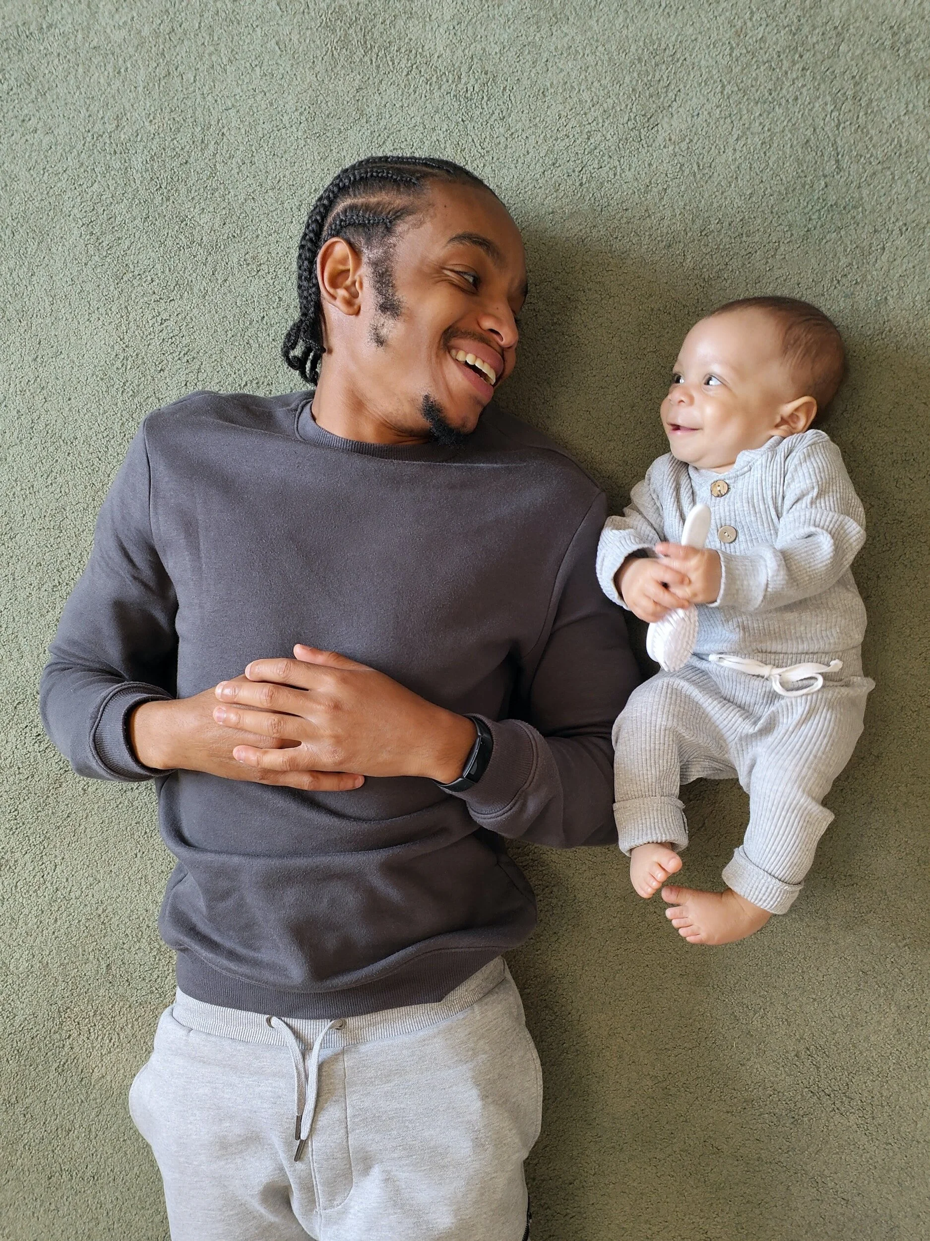 A man and a baby lying on a green carpet looking at each other and smiling.