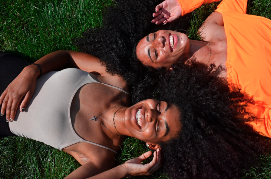 Two women with curly hair lying on grass, smiling and relaxing in the sunlight.